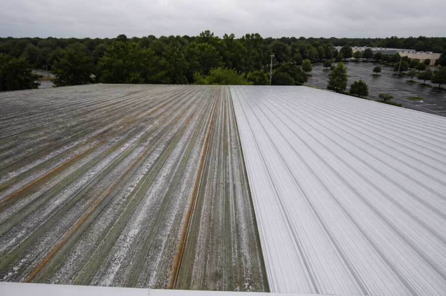 Elevated view of a commercial warehouse in New Jersey with a corrugated metal roof, showing a clear contrast between a weathered section with rust stains, green algae, and oxidation on the left, and a freshly soft-washed, bright clean section on the right. Trees and a mostly empty parking lot are visible in the background under an overcast sky.