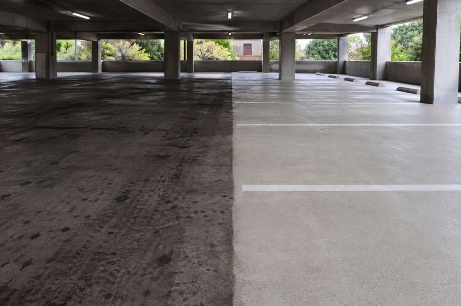 A wide-angle photo inside an empty concrete parking garage in New Jersey showing a clear before-and-after contrast: the left side of the floor is dark with oil stains, tire marks, and grime, while the right side is freshly pressure washed with bright, clean concrete and visible white parking lines, illuminated by overhead fluorescent lights and some natural daylight.