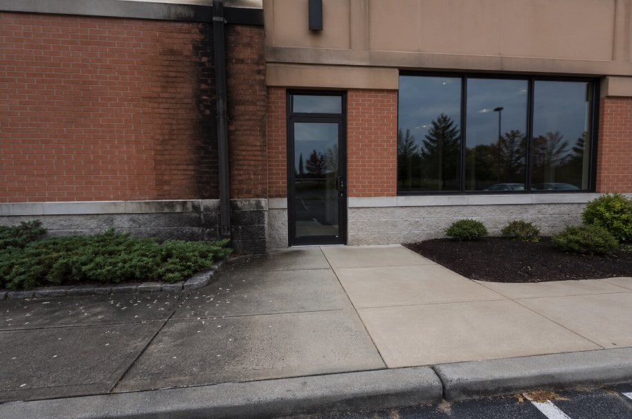 A commercial storefront entrance in a New Jersey shopping plaza, shown in natural daylight. The brick facade is split visually—one side is heavily stained with dark streaks and algae growth, while the other side is clean and bright. The concrete sidewalk below mirrors this contrast, with gum spots and discoloration on the dirty side and a spotless surface on the cleaned side. A glass door and large window sit at the center, with small landscaped beds along the building. No people or signage are visible.