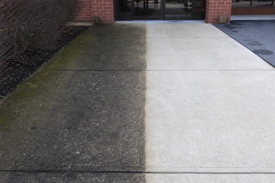 A commercial concrete walkway outside a brick storefront in a New Jersey shopping plaza, shown half dirty with dark stains, gum spots, and green algae along the edge, and half freshly pressure washed, appearing bright and clean, with glass entrance doors in the background and no people present.
